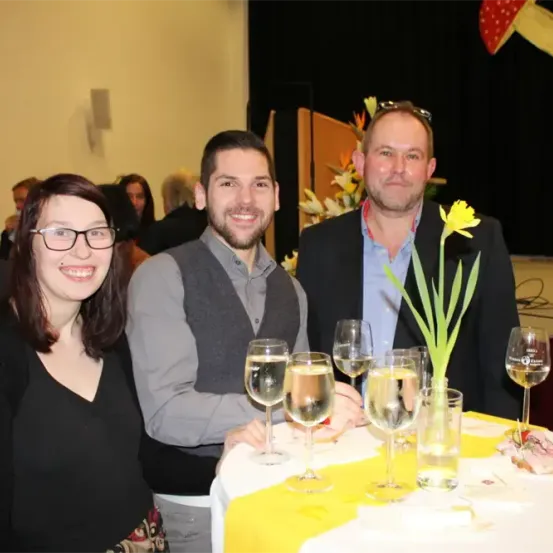 Drei Menschen stehen an einem Tisch und lächeln für ein Foto. Hinter ihnen sind andere verschwommen im Hintergrund. Der Tisch ist mit Gläsern und einer gelben Blume gedeckt.