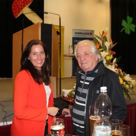 Ein Mann und eine Frau stehen mit Getränken an einem Tisch und lächeln. Dahinter eine Bühne mit Mikrofon und Blumen. Ein Banner mit dem Text 'SPARKASSE'.