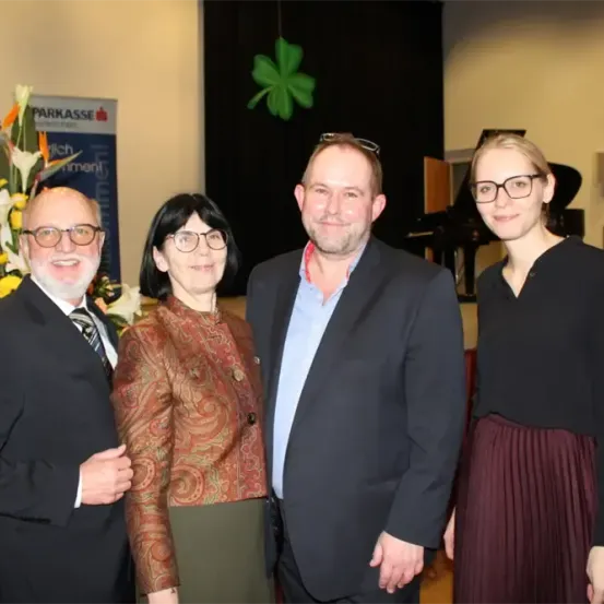 Vier Personen stehen in einem Raum mit einem Klavier im Hintergrund. Der Mann links trägt einen Krawatte, und die anderen sind in formeller Kleidung. Ein großer Blumenstrauß steht hinter ihnen.
