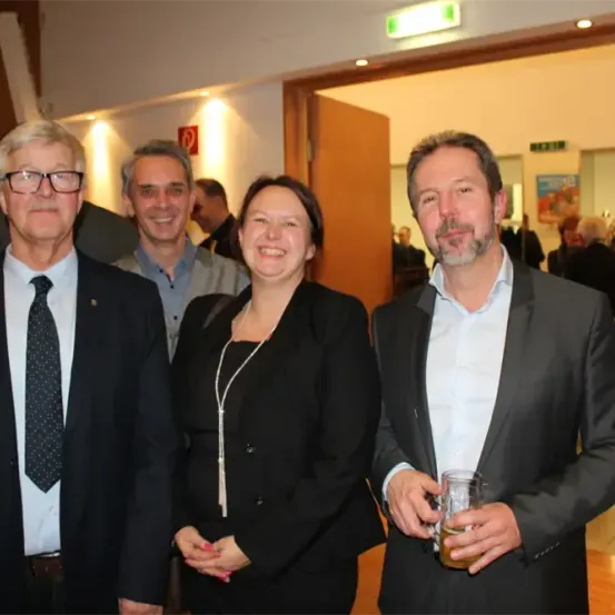 Vier Personen posieren für ein Foto bei einer Veranstaltung. Der Mann auf der linken Seite trägt eine Brille und eine Krawatte. Die Frau in der Mitte lächelt. Der Mann auf der rechten Seite hält ein Glas. Es sind weitere Personen im Hintergrund zu sehen.