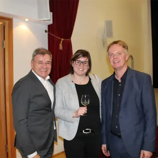 Drei formell gekleidete Personen stehen lächelnd zusammen. Die Frau in der Mitte hält ein Weinglas. Hinter ihnen befindet sich eine Wand mit einem roten Vorhang.