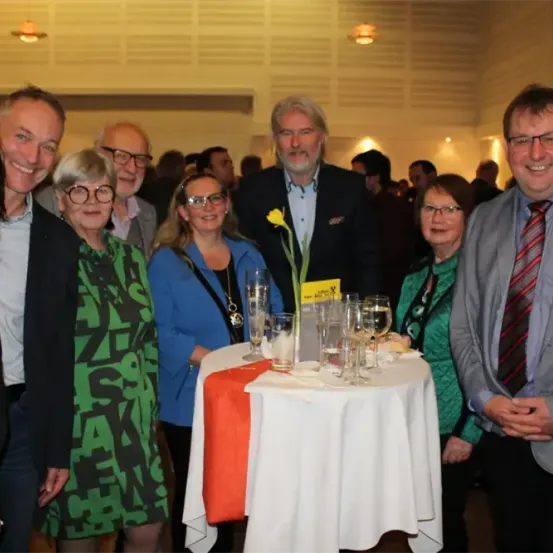 Eine Gruppe von Menschen, die lächeln, versammelt sich um einen Tisch mit Wein glasses und einem Blumen-Centerpiece. Sie scheinen sich auf einer Veranstaltung zu befinden.