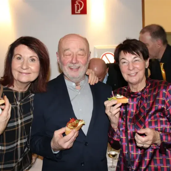 Drei Erwachsene lächeln und halten Essen in den Händen, einer mit einem Sandwich und zwei mit Fingerfood. Ein Mann im Anzug steht in der Mitte. Eine weiße Wand mit einem Feuerlöscher-Schild ist hinter ihnen.