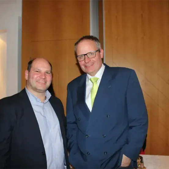 Zwei Männer in Anzügen lächeln für ein Foto. Der Mann auf der rechten Seite trägt eine grüne Krawatte. Hinter ihnen ist eine Holzwand.