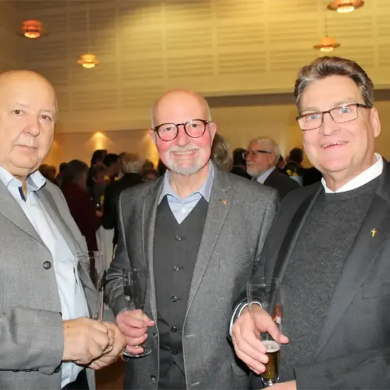 Drei Männer stehen zusammen und halten Gläser in der Hand in einem überfüllten Raum. Der Mann auf der linken Seite trägt einen grauen Anzug. Der Mann in der Mitte trägt ein graues Sakko und eine Brille. Der Mann auf der rechten Seite trägt einen schwarzen Pullover und eine Brille.