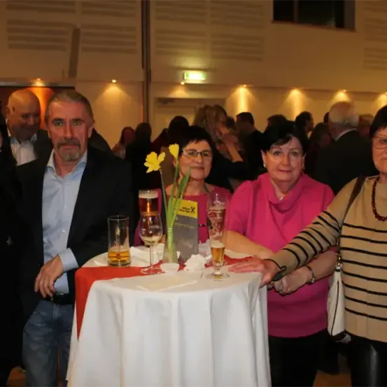 Eine Gruppe von Menschen steht um einen Tisch mit Gläsern und einer Blume, möglicherweise bei einer Veranstaltung. Zwei Frauen tragen Brillen und Halsketten. Dahinter lächeln ein Mann und eine Frau.