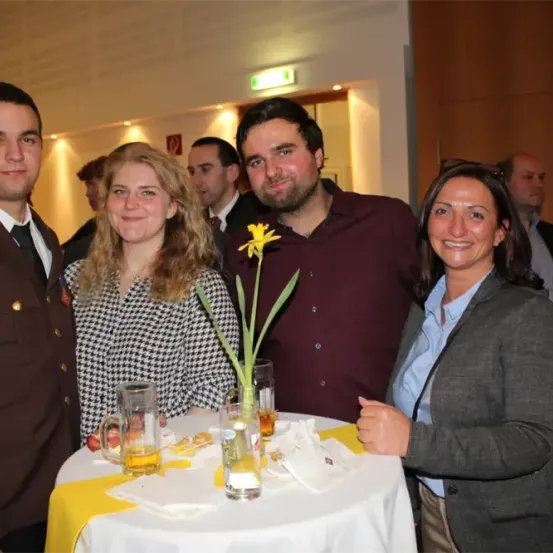 Vier Personen posieren für ein Foto bei einer Zusammenkunft mit einem Tisch, auf dem eine Blume, Gläser und Servietten stehen. Dahinter lächeln ein Mann im Anzug und eine Frau im Anzug.