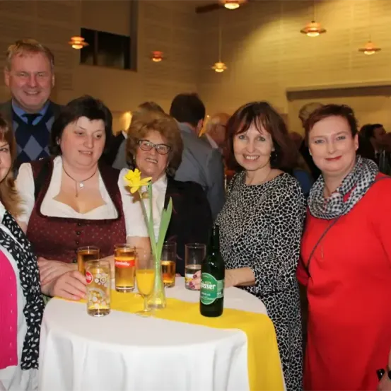 Mehrere Frauen und ein Mann stehen um einen Tisch herum, lächeln und posieren für ein Foto. Der Tisch hat eine gelbe Tischdecke, Gläser und Bierflaschen. Eine gelbe Blume in einer Vase steht ebenfalls auf dem Tisch.