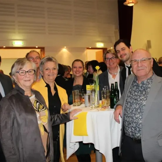 Eine Gruppe von Menschen versammelt sich um einen Tisch bei einer Veranstaltung und lächelt für ein Foto. Auf dem Tisch stehen Gläser, Flaschen und eine Blume. Sie sind feierlich gekleidet.