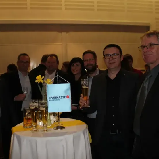 Eine Gruppe von Menschen steht um einen Tisch mit Biergläsern, hält Getränke in der Hand, lächelt und posiert für ein Foto. Der Tisch hat ein Banner mit dem Text 'Sparkasse Neunkirchen'.