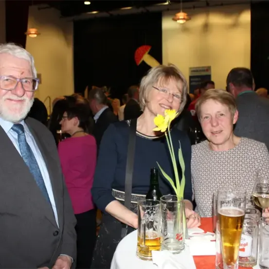 Drei Personen posieren an einem Tisch mit Getränken. Eine Frau hält eine Blume und lächelt. Der Mann links trägt einen Anzug und eine Brille. Der Tisch ist mit Gläsern, Flaschen und Servietten geschmückt.