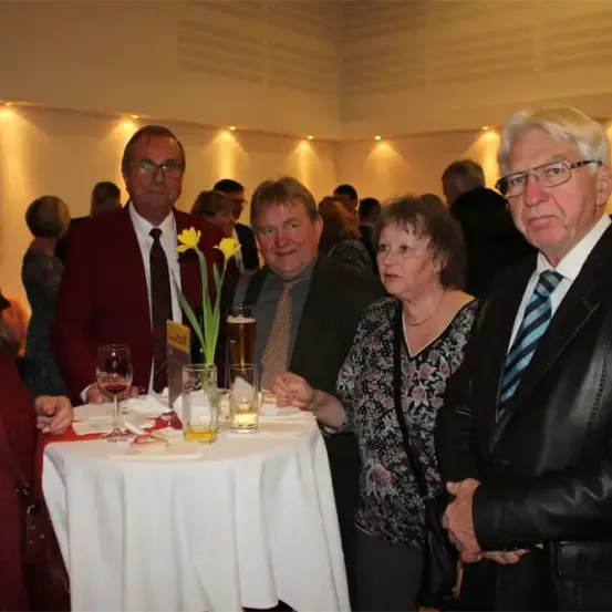 Eine Gruppe von Menschen steht um einen Tisch mit einer weißen Tischdecke herum. Sie scheinen eine formelle Veranstaltung zu besuchen. Der Tisch hat eine Blumenvase, ein Weinglas und eine Tasse. Die Frau auf der linken Seite hält ein Weinglas. Der Mann in der Mitte trägt eine Krawatte und eine Brille. Der Mann auf der rechten Seite trägt eine Lederjacke.