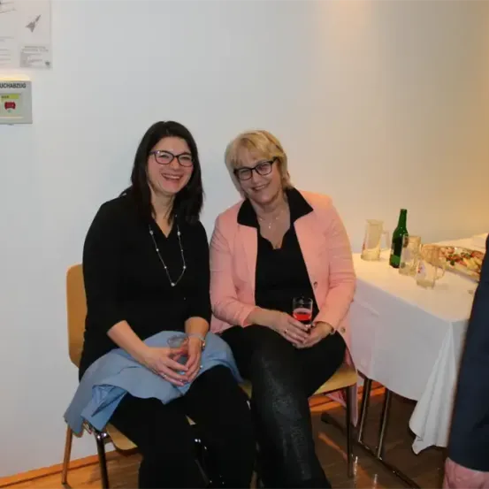 Zwei Frauen in Brillen sitzen lächelnd nebeneinander, mit einem Tisch und Getränken dahinter, in der Nähe einer weißen Wand mit einem Rauchverbotsschild.