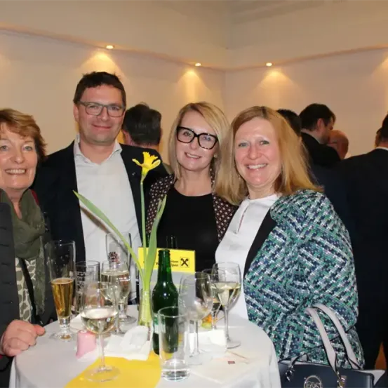 Drei Frauen und ein Mann stehen um einen Tisch mit Weingläsern und einer gelben Blume. Der Mann trägt eine Brille und einen Anzug. Sie sind bei einer Veranstaltung, und es gibt Leute in Anzügen im Hintergrund.