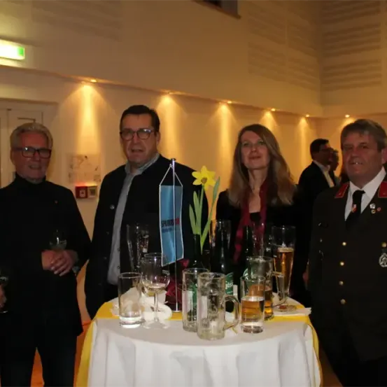 Fünf Personen stehen um einen Tisch mit Gläsern und Flaschen, eine Frau hält eine gelbe Blume. Sie scheinen sich auf einer formellen Veranstaltung zu befinden.