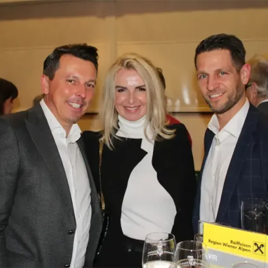 Drei Menschen stehen lächelnd vor einem gelben Schild. Ein Mann und eine Frau tragen formelle Kleidung. Hinter ihnen sind ein Mann mit Bart und eine Frau mit grauen Haaren teilweise sichtbar.