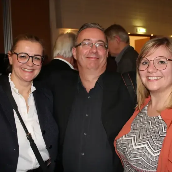 Drei Menschen stehen eng beieinander und lächeln in die Kamera. Zwei Frauen und ein Mann. Sie tragen Brillen. Der Hintergrund ist mit Menschen verwischt.