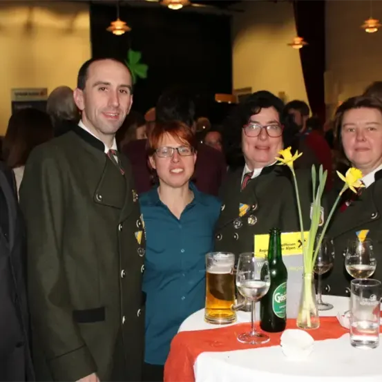 Fünf Personen, drei in Uniform, stehen lächelnd bei einer Veranstaltung. Eine Frau in einem blauen Shirt hält Blumen, und ein Tisch mit Getränken und einer grünen Flasche ist im Vordergrund.