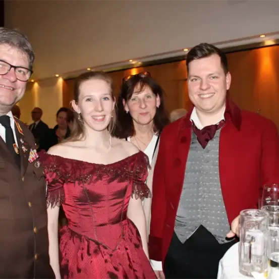 Eine Gruppe von Menschen in formeller Kleidung steht bei einer Veranstaltung zusammen, lächelt und posiert für ein Foto. Im Vordergrund befinden sich Gläser und eine Flasche auf einem Tisch.