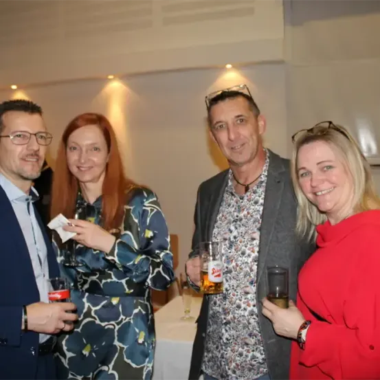 Vier Menschen lächeln und posieren für ein Foto, zwei Männer und zwei Frauen, mit Getränken in der Hand, wahrscheinlich bei einer gesellschaftlichen Veranstaltung.