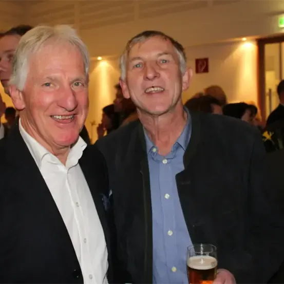 Zwei Männer lächeln für ein Foto auf einer Party. Einer hält ein Glas Bier. Viele Menschen sind im Hintergrund verschwommen.