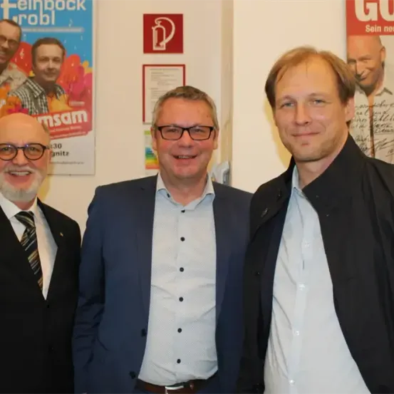 Drei Männer stehen nebeneinander vor einer Wand mit Postern. Sie alle lächeln und scheinen für ein Foto zu posieren.