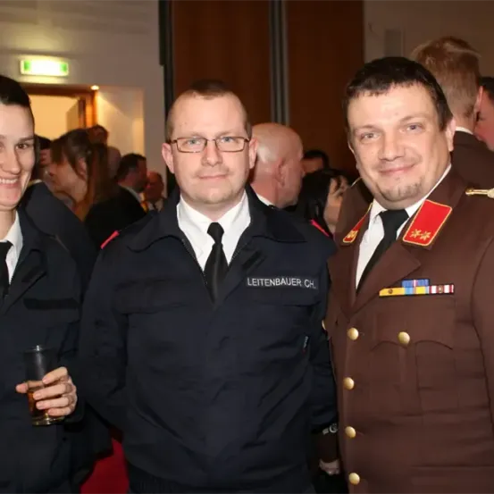 Drei uniformierte Personen, zwei Männer und eine Frau, stehen bei einer Veranstaltung zusammen und lächeln. Der Mann rechts trägt eine braune Militäruniform mit Medaillen.