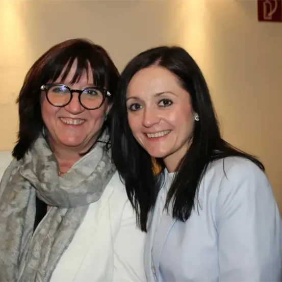 Zwei Frauen, eine mit Brille und Schal, die andere mit Ohrringen, lächeln und posieren für ein Foto in einem Innenraum.