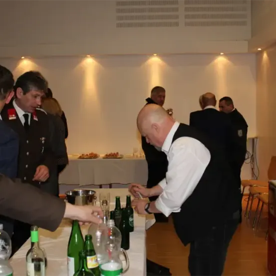 Ein Mann in einem schwarzen Sakko öffnet eine Flasche, während er an einem Tisch steht, an dem sich mehrere andere in formeller Kleidung befinden. Es scheint sich um eine gesellschaftliche Zusammenkunft oder Veranstaltung zu handeln.