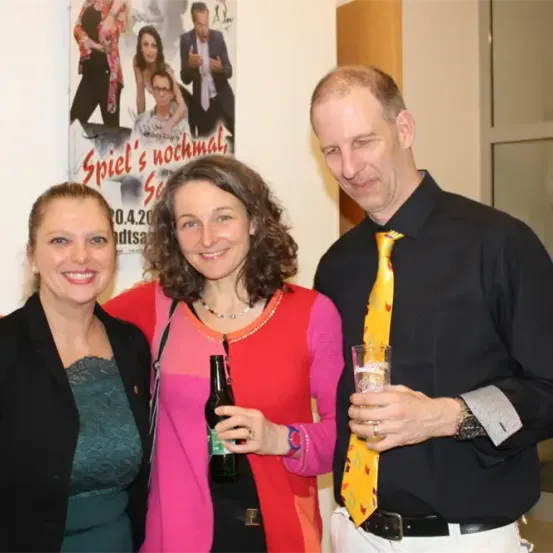 Drei Menschen stehen nebeneinander und lächeln für ein Bild. Eine Frau hält eine Flasche und ein Mann ein Glas. Sie stehen vor einem Poster.