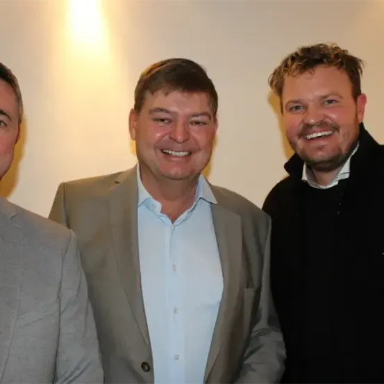Drei Männer stehen eng beieinander und lächeln für ein Foto. Der Mann auf der linken Seite trägt einen grauen Anzug. Der Mann in der Mitte trägt einen hellgrauen Anzug und ein blaues Hemd. Der Mann auf der rechten Seite trägt einen schwarzen Mantel.