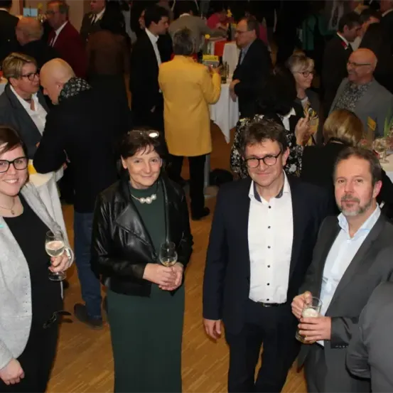 Eine Gruppe von Menschen steht zusammen, wahrscheinlich bei einer formellen Veranstaltung. Vier Personen sind im Vordergrund zu sehen, lächelnd und mit Gläsern in der Hand. Der Holzboden deutet auf eine elegante Atmosphäre hin.