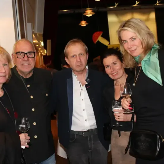 Fünf Personen, darunter eine Frau mit einem grünen Schal, stehen zusammen, lächeln und halten Weingläser. Der Mann in der Mitte trägt ein weißes Hemd, die anderen sind in Schwarz gekleidet. Sie scheinen sich bei einer gesellschaftlichen Veranstaltung zu befinden.