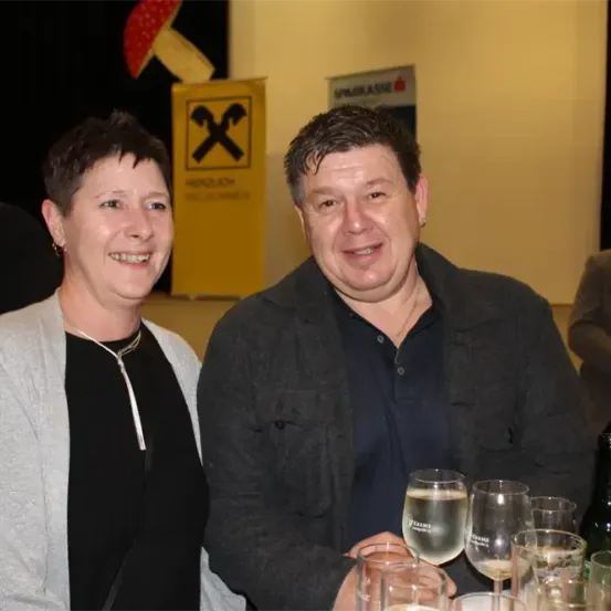 Ein Mann und eine Frau stehen lächelnd neben einem Tisch mit Weingläsern und Flaschen. Der Mann trägt eine Halskette. Ein gelbes Schild mit einem 'X' und ein roter Pilz sind hinter ihnen.