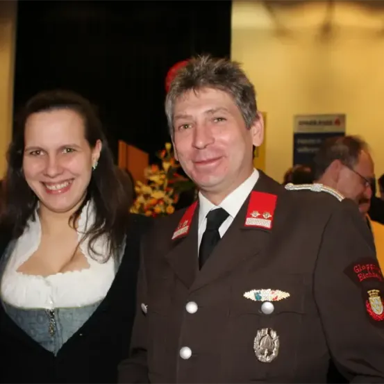 Ein Mann in Uniform und eine Frau in traditioneller Kleidung stehen lächelnd nebeneinander vor der Kamera. Dahinter ist eine Menschenmenge undeutlich zu sehen, sowie ein Blumenarrangement und ein Banner im Hintergrund.