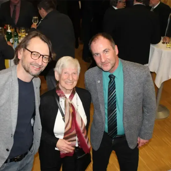 Drei Personen stehen bei einer formellen Veranstaltung zusammen. Der Mann links trägt eine Brille und eine graue Jacke, die Frau in der Mitte trägt einen Schal und einen schwarzen Blazer, und der Mann rechts trägt einen grauen Blazer und eine Krawatte. Dahinter steht ein Tisch mit weißer Tischdecke und Blumenarrangement.