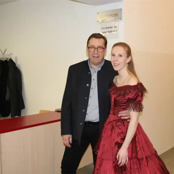Ein Mann und eine Frau stehen eng nebeneinander vor einer Wand und lächeln für ein Foto. Der Mann trägt eine Brille und einen schwarzen Anzug. Die Frau trägt ein rotes Kleid. Hinter ihnen befindet sich eine weiße Wand mit einem Schild, das 'Garderobe' besagt.