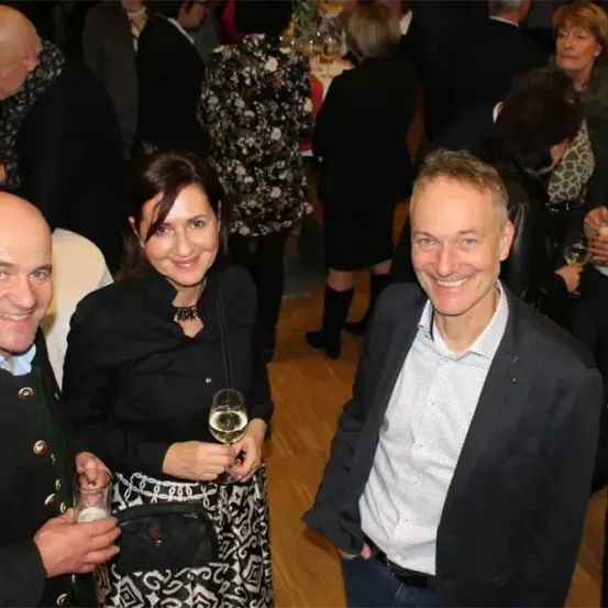 Drei Menschen lächeln bei einer gesellschaftlichen Veranstaltung. Eine Frau hält ein Weinglas und ein Mann neben ihr hält ein Getränk in der Hand. Hinter ihnen unterhalten sich mehrere Menschen.