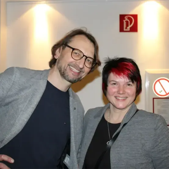 Ein Mann und eine Frau stehen gemeinsam vor einer weißen Wand. Der Mann trägt eine Brille und eine graue Jacke, und die Frau hat rote Haare und trägt eine graue Jacke. Sie lächeln beide für die Kamera. Hinter ihnen befindet sich ein Feuerlöscher-Schild und ein Rauchverbotsschild.