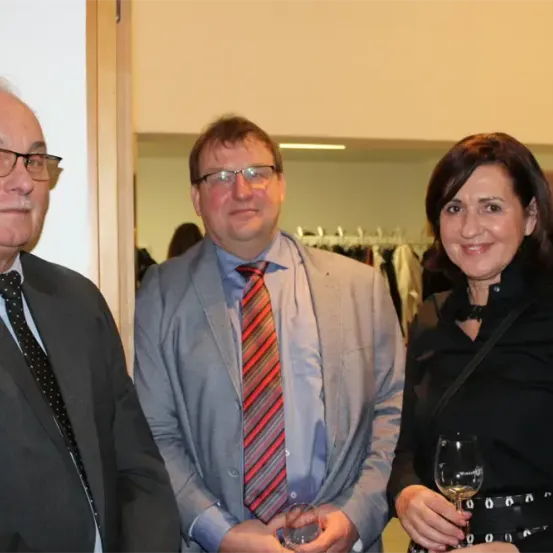 Drei formell gekleidete Menschen, zwei Männer und eine Frau, stehen zusammen. Der Mann auf der linken Seite trägt einen schwarzen Anzug und eine Krawatte. Der Mann in der Mitte trägt einen grauen Anzug und eine gestreifte Krawatte. Die Frau auf der rechten Seite trägt ein schwarzes Kleid und hält ein Weinglas. Sie stehen vor einer weißen Wand.