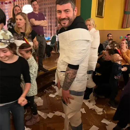 Ein Mann, der in Toilettenpapier eingewickelt ist, steht unter einer Gruppe von Menschen in einem festlichen Ambiente, wobei einige lachen und andere zusehen. Papiere sind auf dem Boden verstreut.
