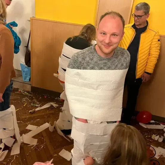 Ein in Toilettenpapier eingewickelter Mann steht in einem unordentlichen Raum mit anderen, einige schauen ihn an. Eine Frau mit einem Pferdeschwanz schaut ihn an. Ein Mann in einer gelben Jacke ist in der Nähe.