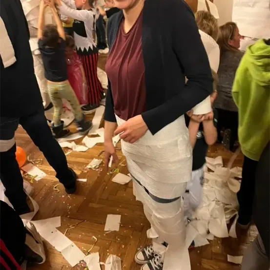 Eine in Toilettenpapier eingewickelte Frau steht in einem Raum mit verstreutem Papier, umgeben von Menschen und Kindern.