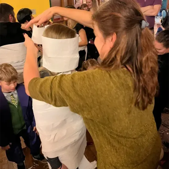 Eine Frau wickelt Toilettenpapier um den Kopf eines Kindes, während andere zusehen. Das Kind hat eine Papierblinde.