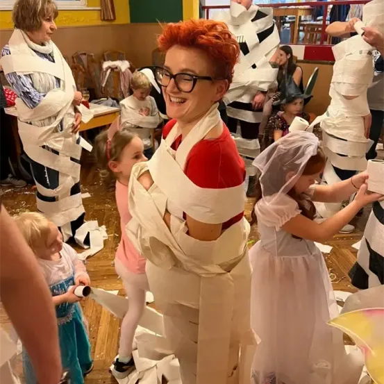 Eine Frau mit roten Haaren und Brille ist in Toilettenpapier eingewickelt, umgeben von Kindern, einer hält eine Rolle Papier. Der Raum ist mit Menschen gefüllt, einige sind ebenfalls in Toilettenpapier eingewickelt.