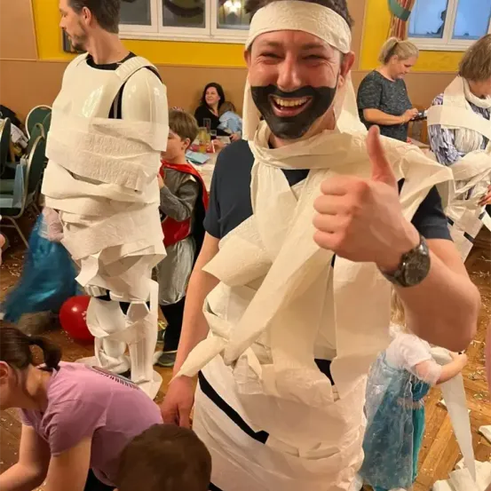 Ein Mann in einem weißen Kostüm, das mit Toilettenpapier bedeckt ist, gibt einen Daumen hoch. Im Hintergrund sind andere Menschen in Kostümen zu sehen. Eine Frau und ein Kind sind ebenfalls anwesend.