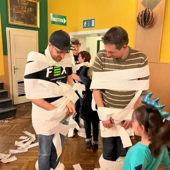 Mehrere Menschen sind in einem Raum, zwei Männer sind in Toilettenpapier eingewickelt, eine Frau beobachtet sie, ein Kind schaut zu und ein Schild sagt 'Toilette.'
