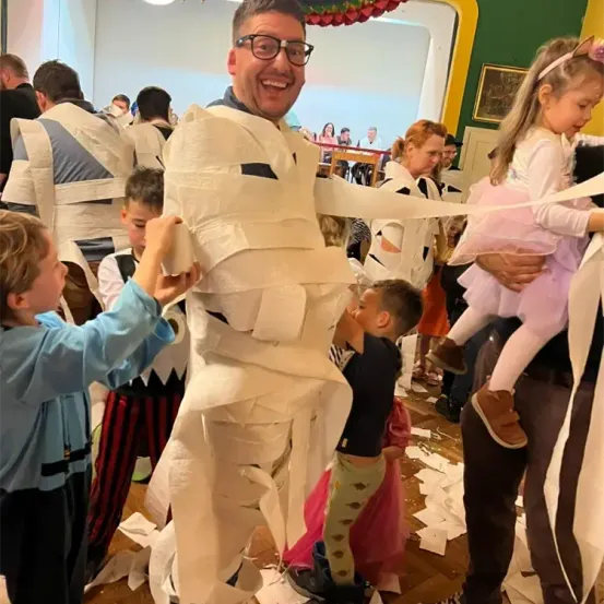 Ein Mann in einem Anzug aus Toilettenpapier lächelt, während Kinder ihn umgeben und Papierstücke halten. Ein Mädchen in einem rosa Kleid hält ein Stück Papier in der Hand.