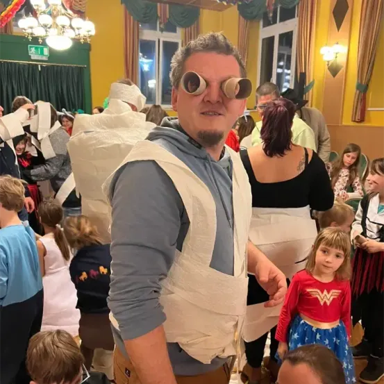 Ein Mann mit einem selbstgemachten Kostüm und Papierbrille steht in einer Menschenmenge von Kindern und Erwachsenen, einige in Kostümen, in einem dekorierten Raum mit einem Kronleuchter und Vorhängen.