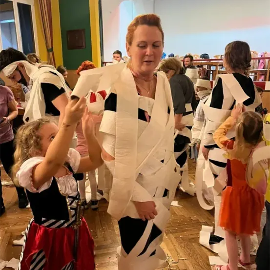 Eine in Toilettenpapier gewickelte Frau steht mit einem kleinen Mädchen. Sie sind in einem Raum mit anderen Leuten, einige sind ebenfalls in Toilettenpapier gewickelt.